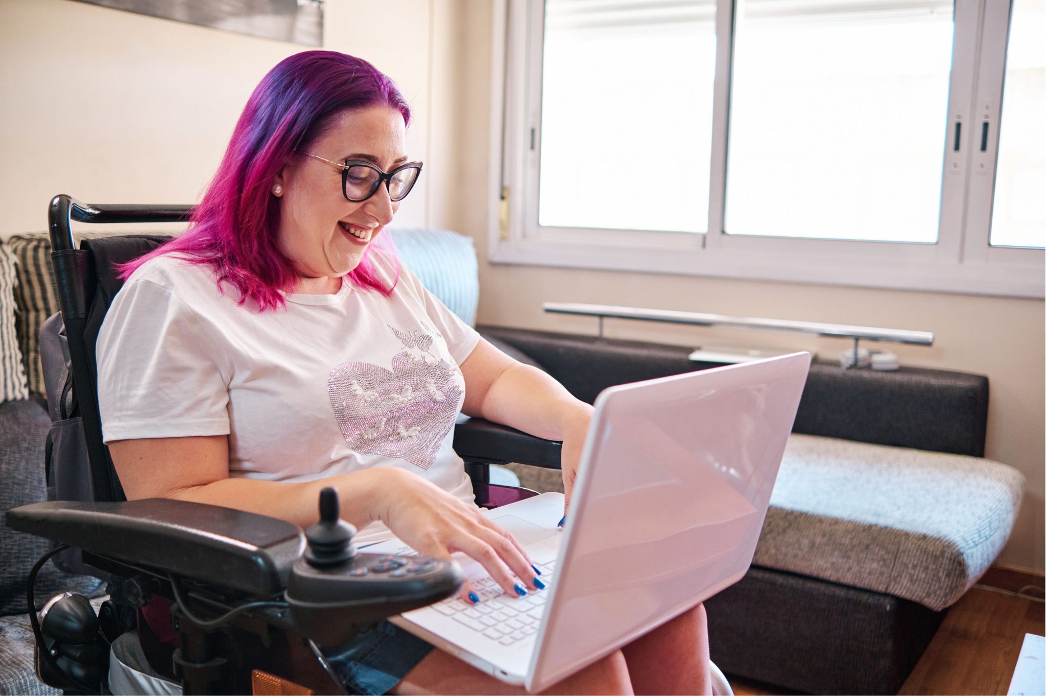 A woman with purple hair and glasses sits in a wheelchair, smiling as she types on a laptop in a cozy living room.
