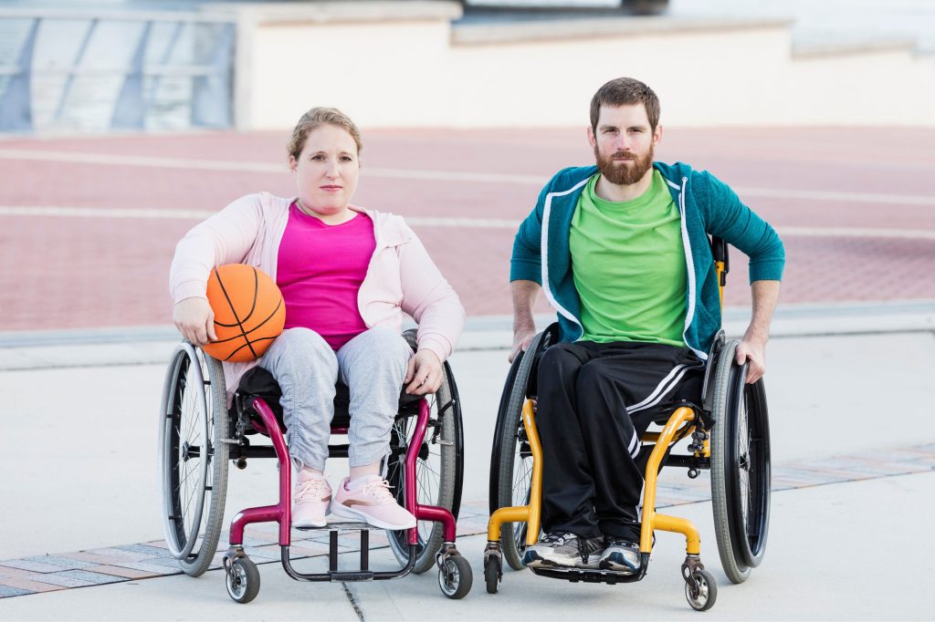 A woman and a man in wheelchairs pose outdoors as she holds a basketball.