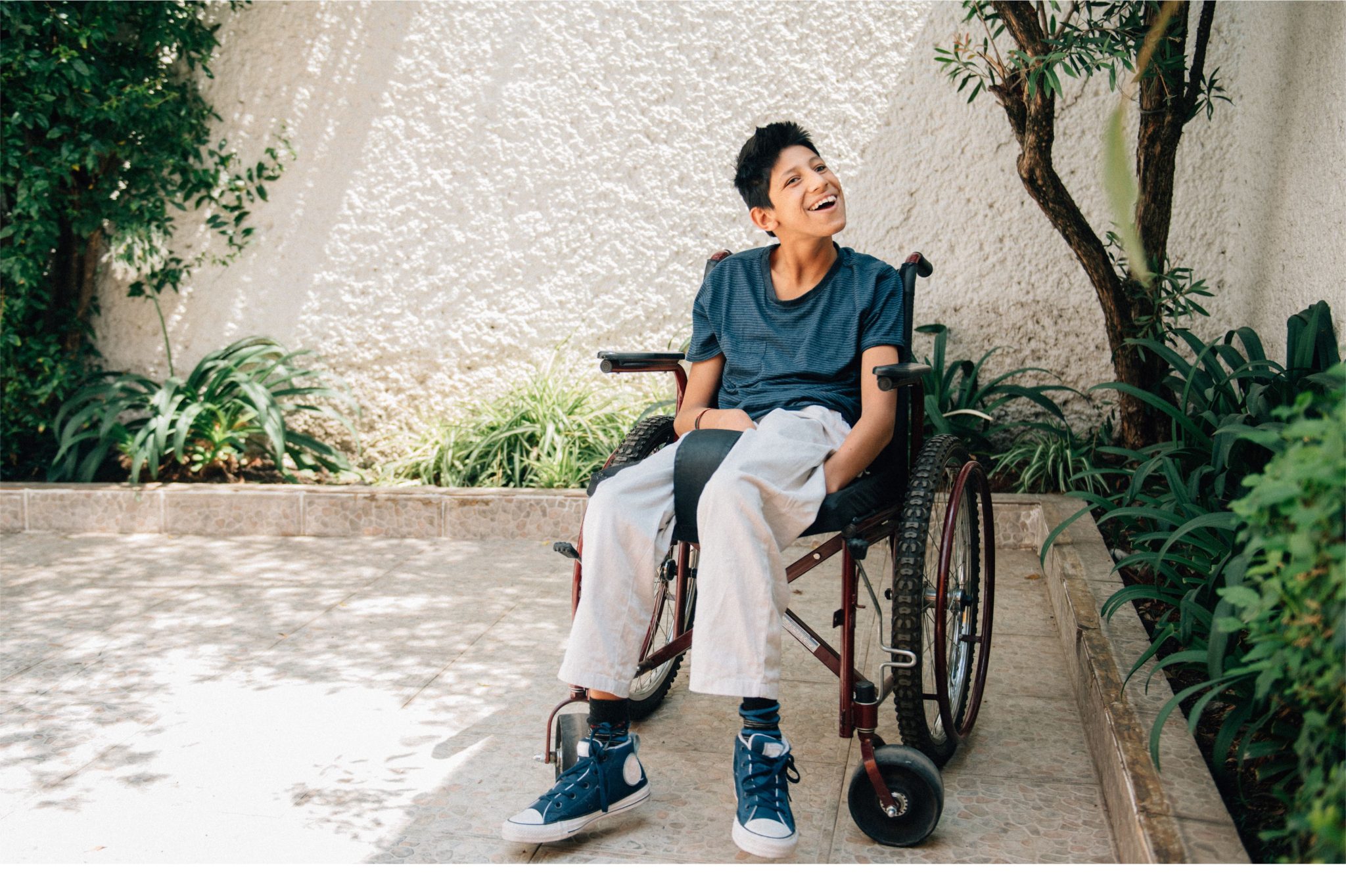 A young boy seated on a wheelchair in a sunlit garden, surrounded by lush greenery against a textured wall.