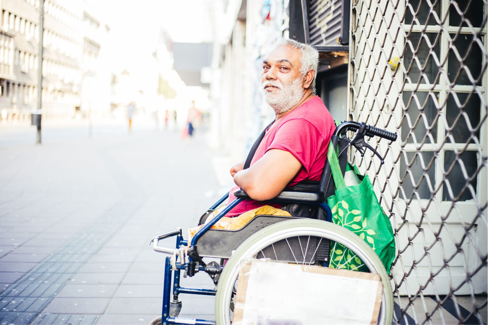 Elderly man with a beard and thoughtful expression sits in a wheelchair on a sunlit city street. He's wearing a pink shirt and has a green bag.