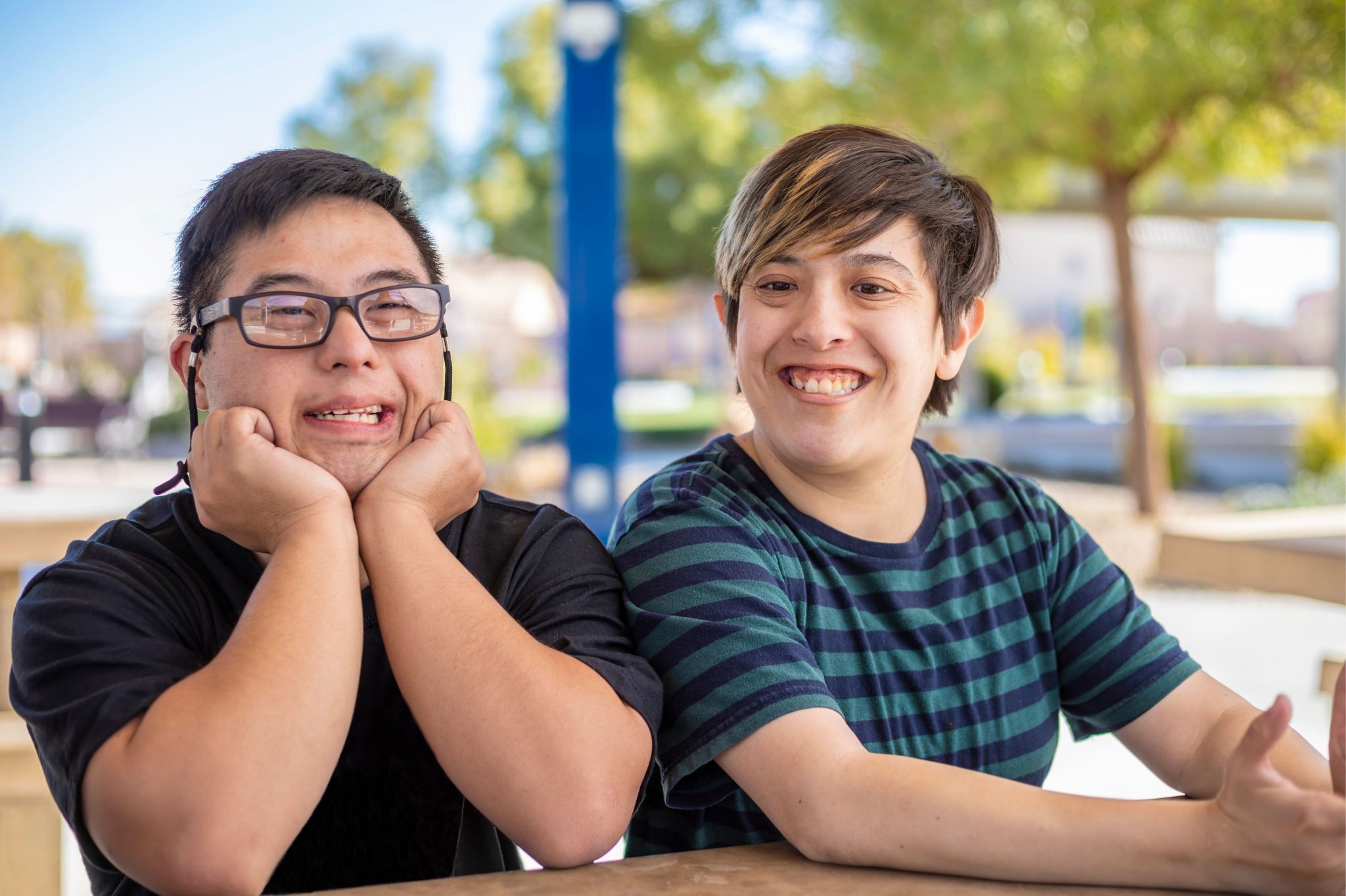 Two friends seated beside each other at a park, smiling and enjoying a sunny day. One wears glasses and rests their chin on hands.