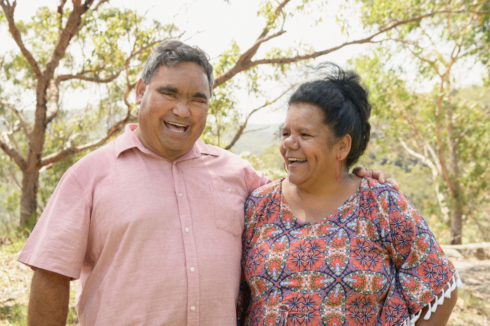 A smiling couple stands together outdoors, surrounded by trees.