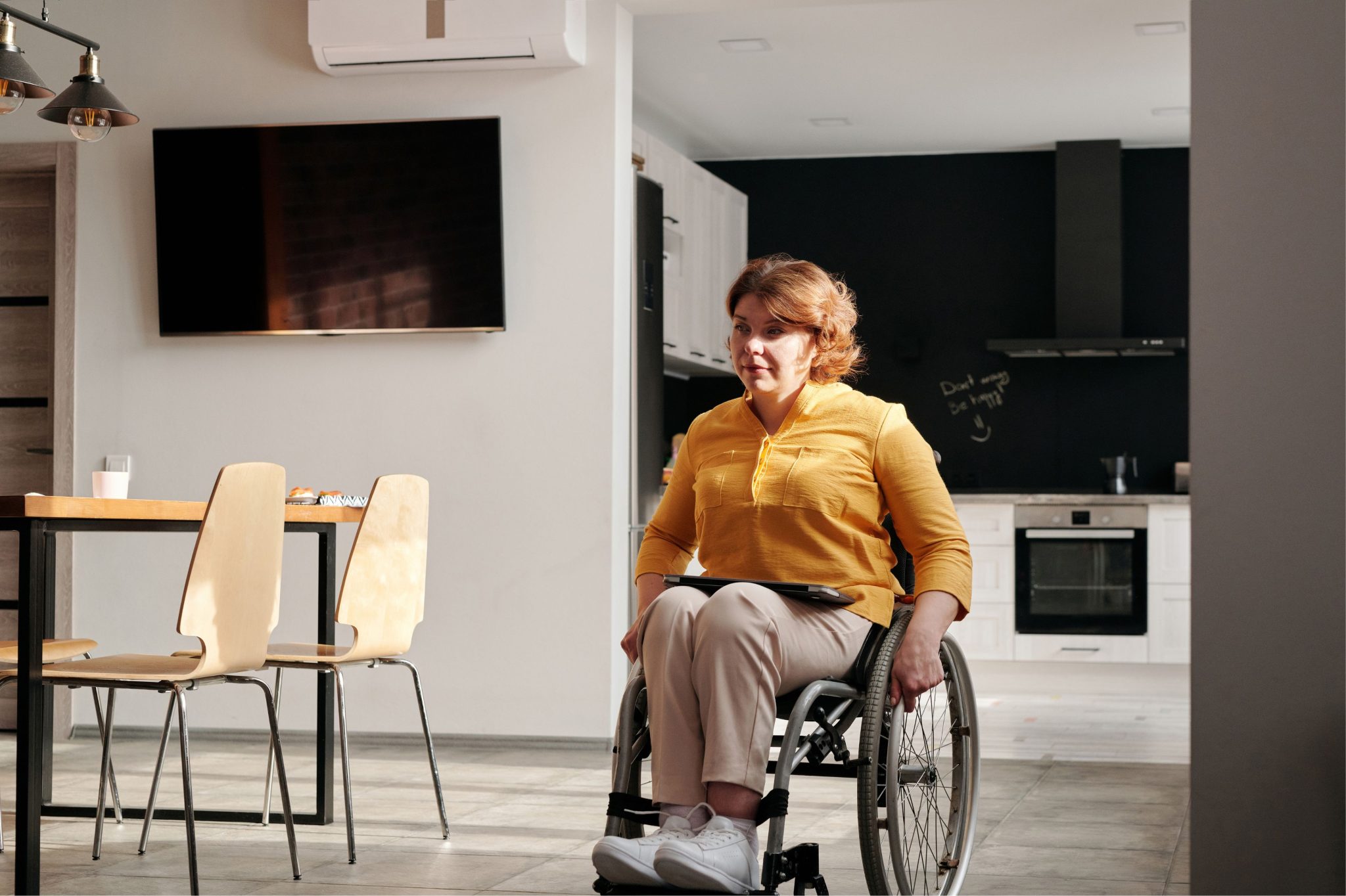 A woman in a yellow sweater sits in a wheelchair in her kitchen with a dining table and chairs.