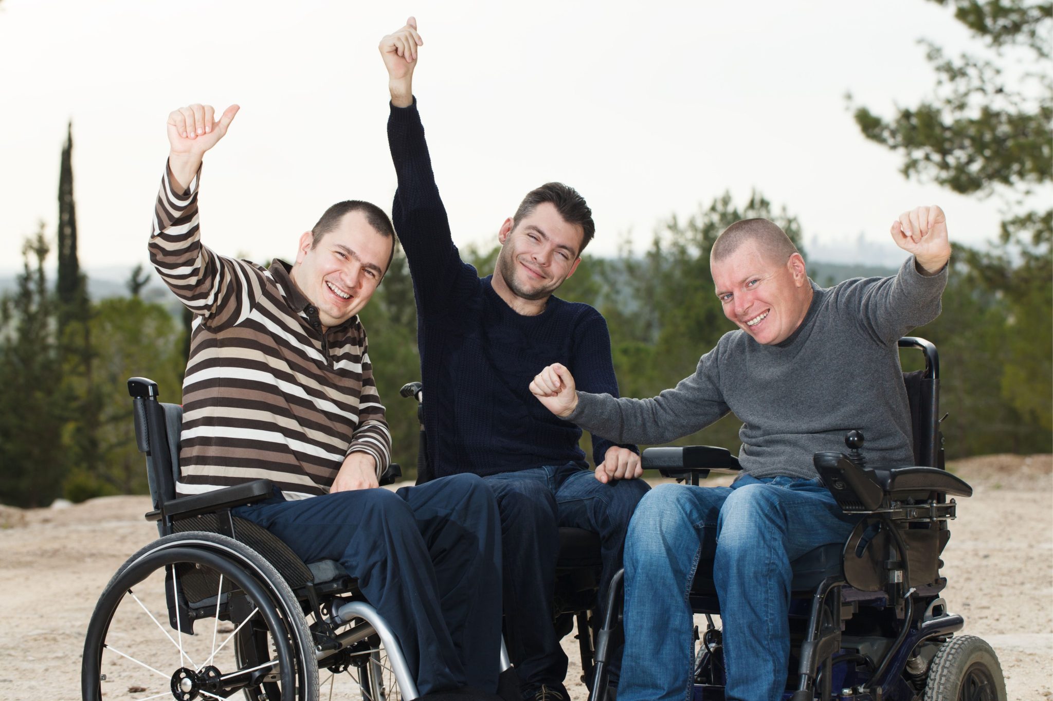 3 men seated on wheelchairs smiling with their arms raised and a thumbs up.