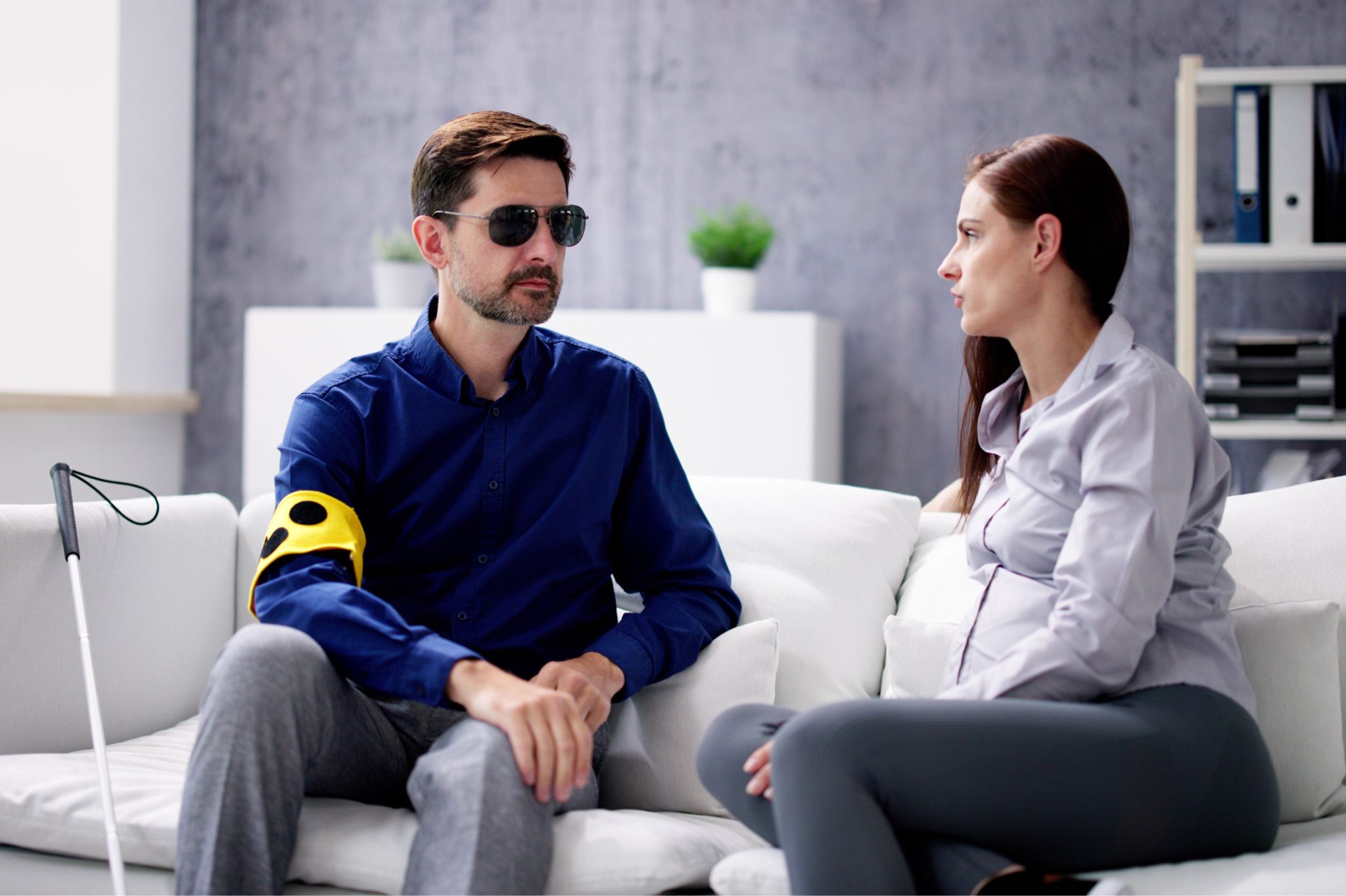 A blind man and a woman seated ona sofa, speaking to each other.