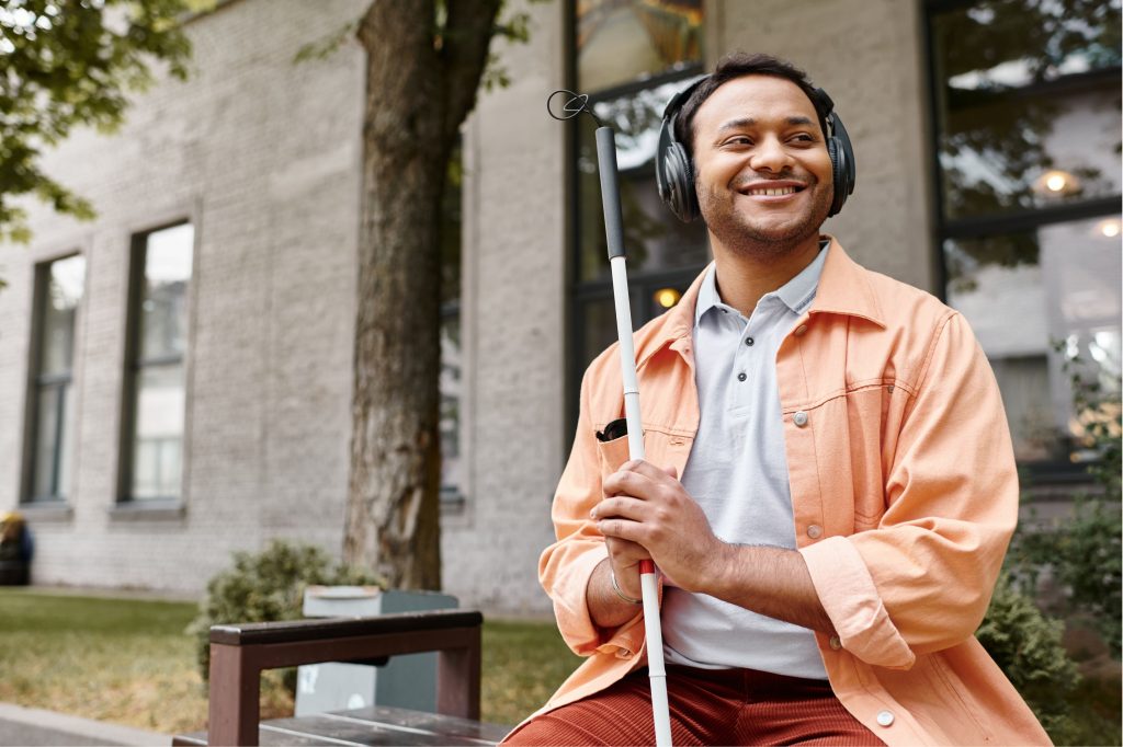 A smiling man, wearing headphones and holding a white support cane, sits on a bench outdoors with a building in the background.