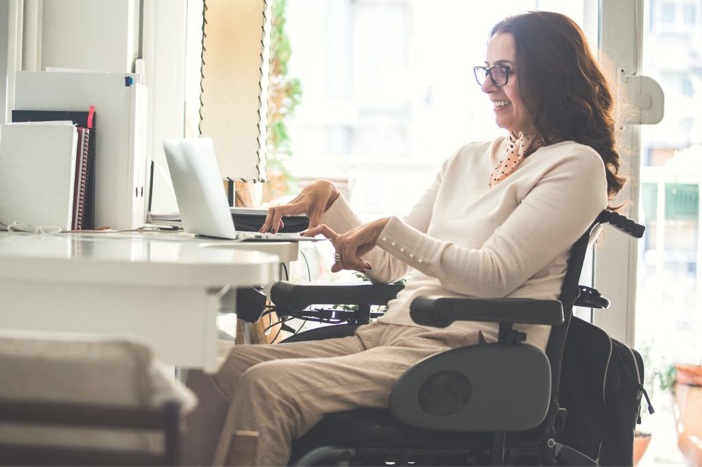 A woman sitting on a wheelchair at her desk typing on a laptop.