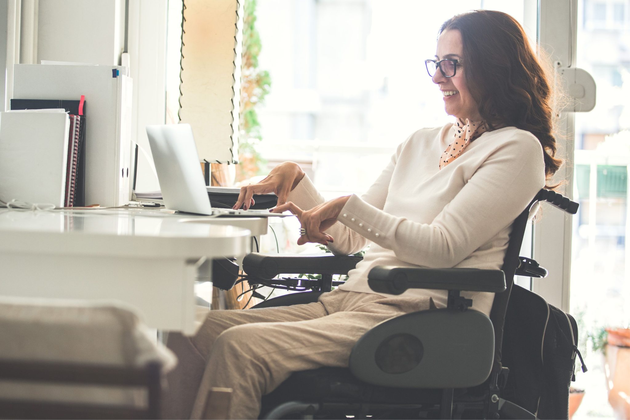 A woman sitting on a wheelchair at her desk typing on a laptop.