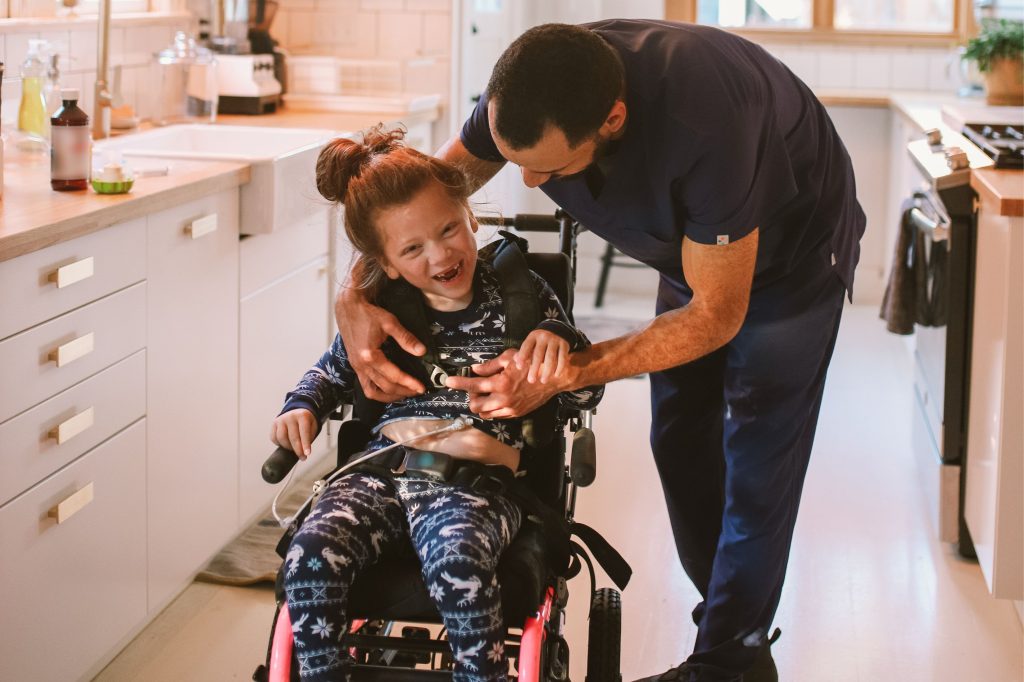 A caregiver in blue gently interacts with a smiling child in a wheelchair in a cozy kitchen.
