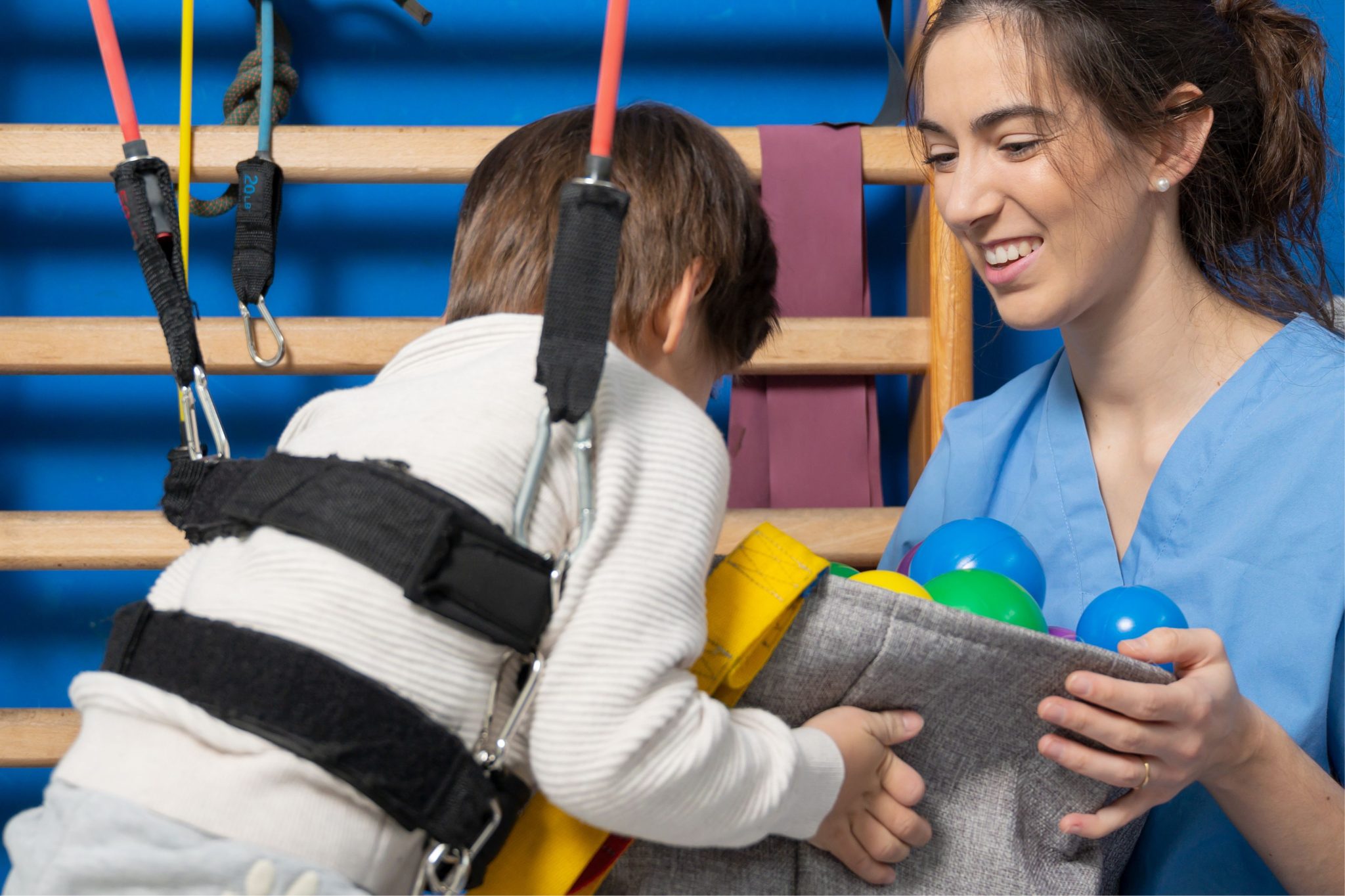 A child in a harness reaches for colorful balls held by a smiling carer in a blue uniform.