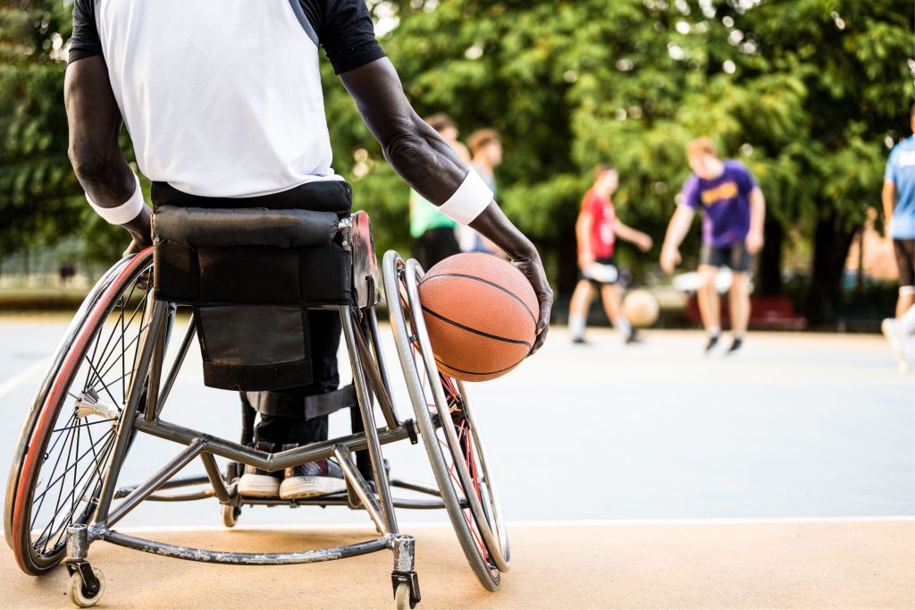 A person in a wheelchair holding a basketball on a court, with a blurred group playing in the background - representing inclusivity and determination.