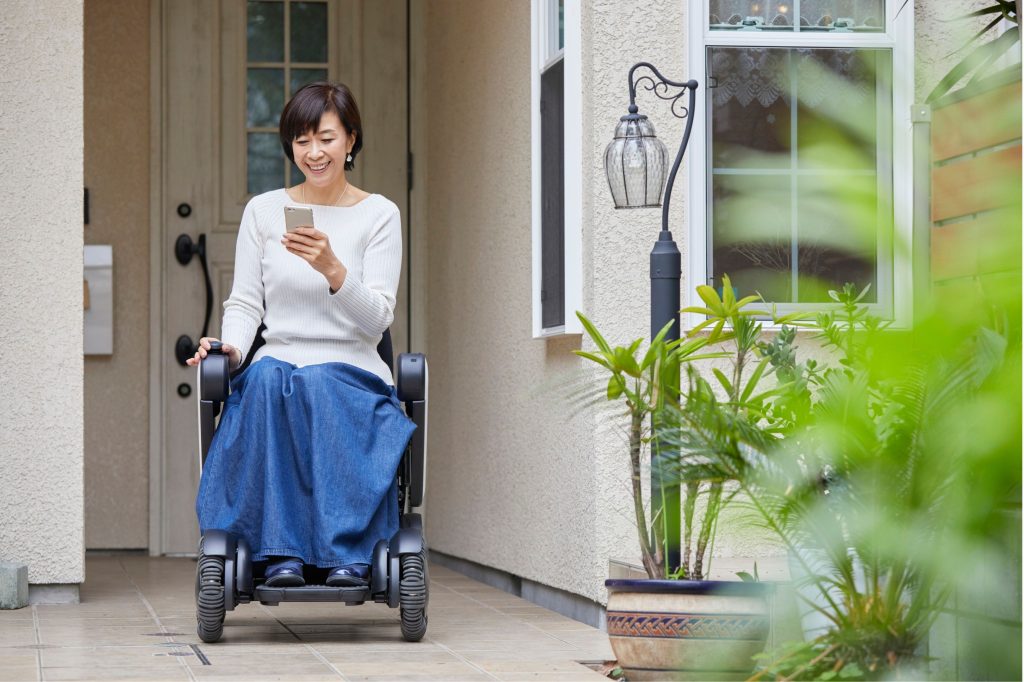 A woman seated on a wheelchair in front of a house, looking at her phone.