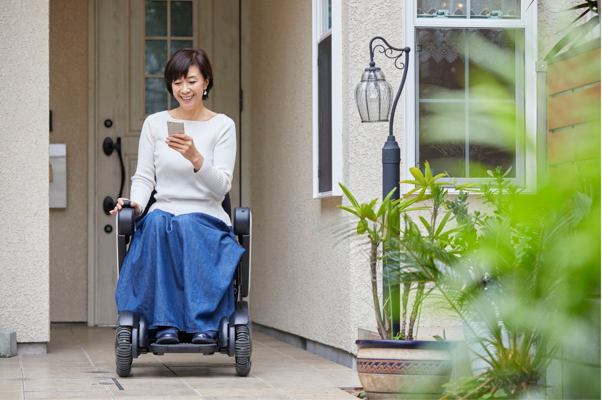 A woman seated on a wheelchair in front of a house, looking at her phone.