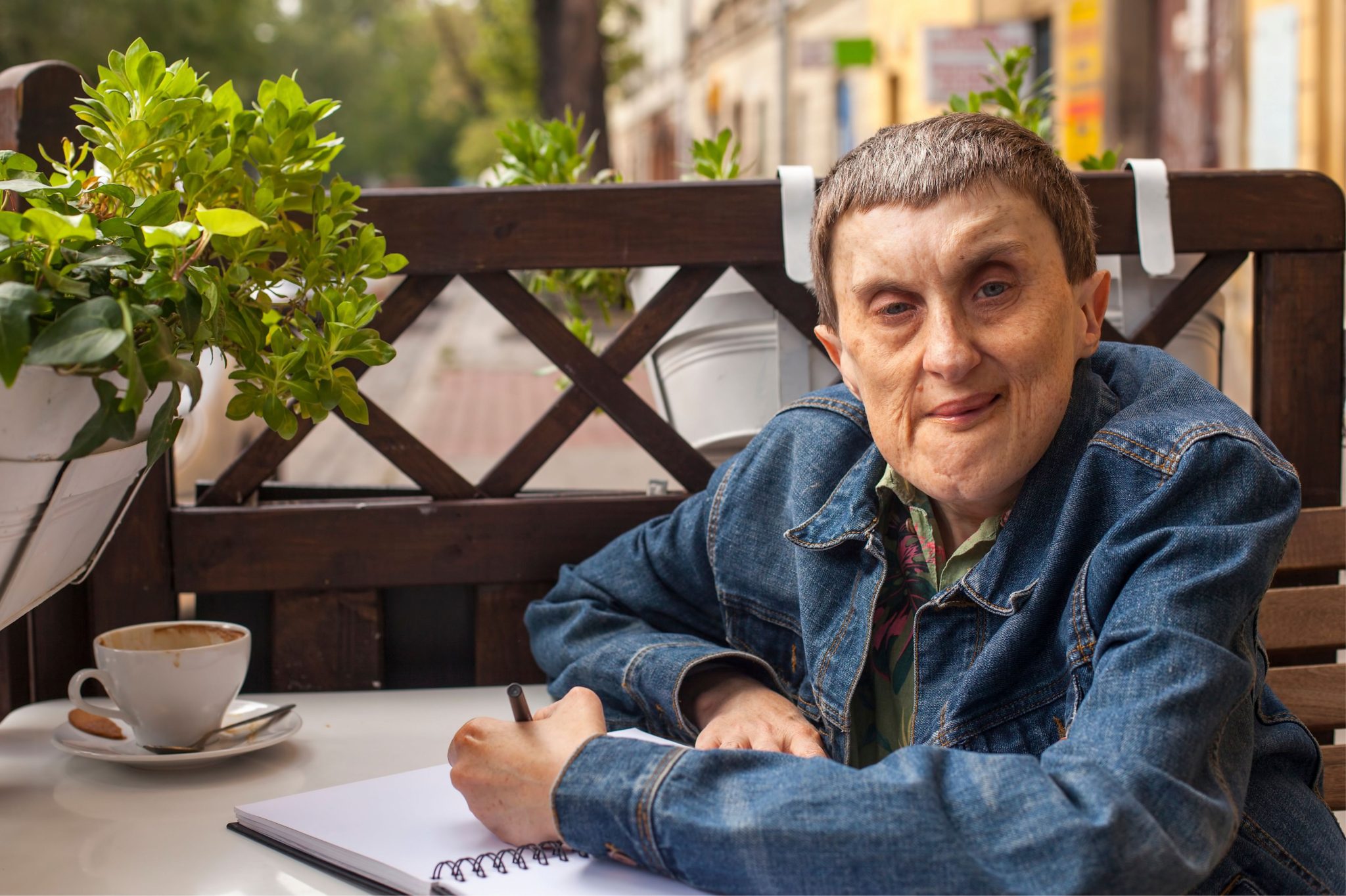 A person in a denim jacket sits at a café table with a cup of coffee, writing in a notebook. Green plants and a wooden fence nearbycreate a cozy atmosphere.