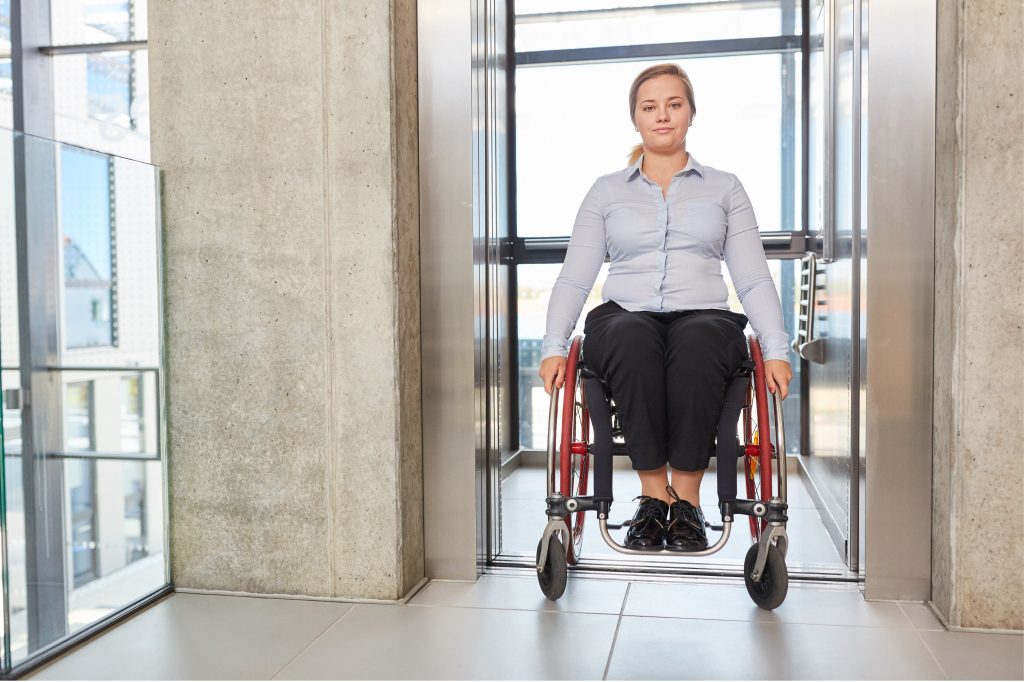 Woman wearing a light blue shirt and black pants, seated on a wheelchair exiting from an elevator in a modern building.