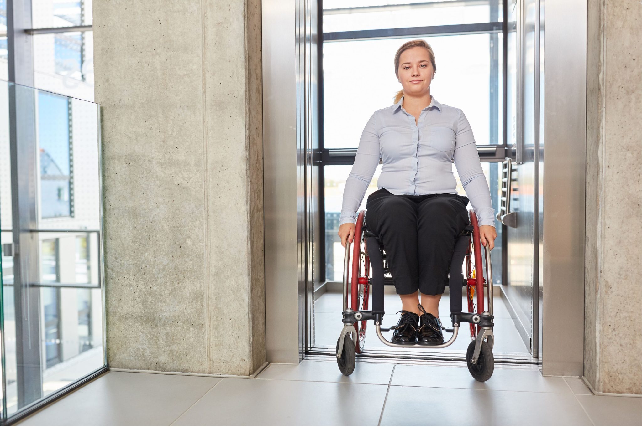 Woman wearing a light blue shirt and black pants, seated on a wheelchair exiting from an elevator in a modern building.