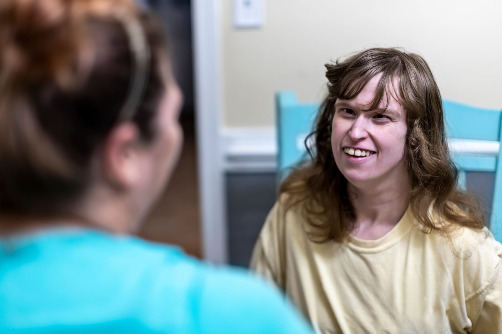 A woman in a yellow shirt, seated on a blue chair, smiles warmly at another person sitting facing her. The setting looks relaxed and friendly.