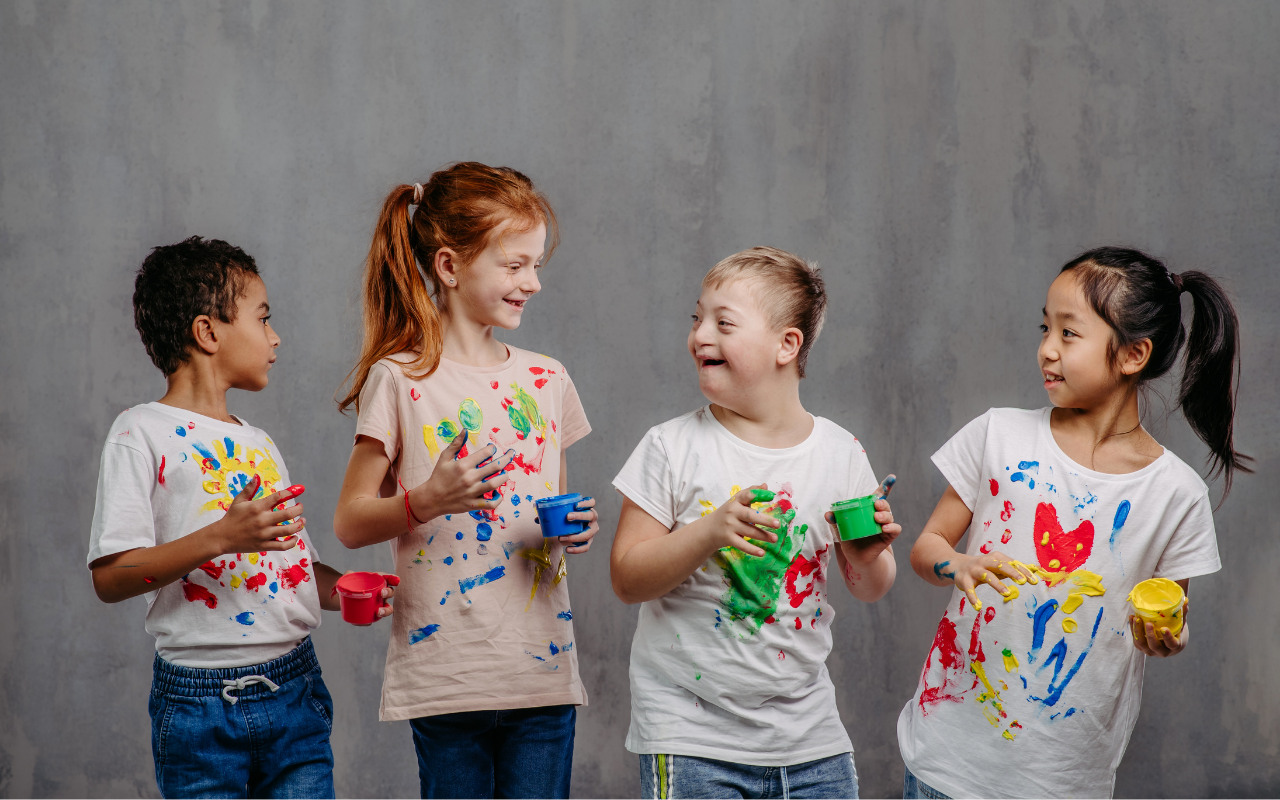 Four children standing against a gray background smile and hold paint containers, wearing white t-shirts with colorful paint splatters.